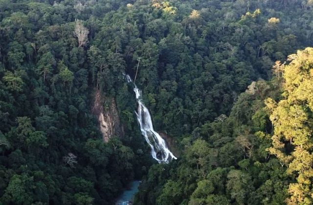 Lapopu Waterfall cascades down a steep, dark green forested cliff into a light blue river below, surrounded by dense tropical trees.