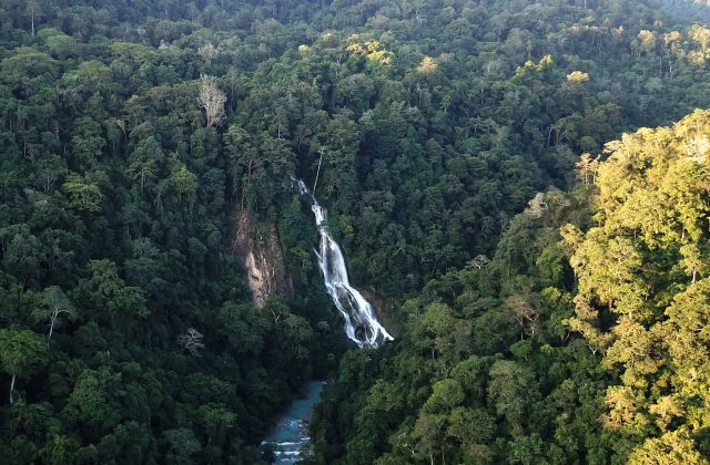 Lapopu Waterfall cascades down a steep, dark green forested cliff into a light blue river below, surrounded by dense tropical trees.