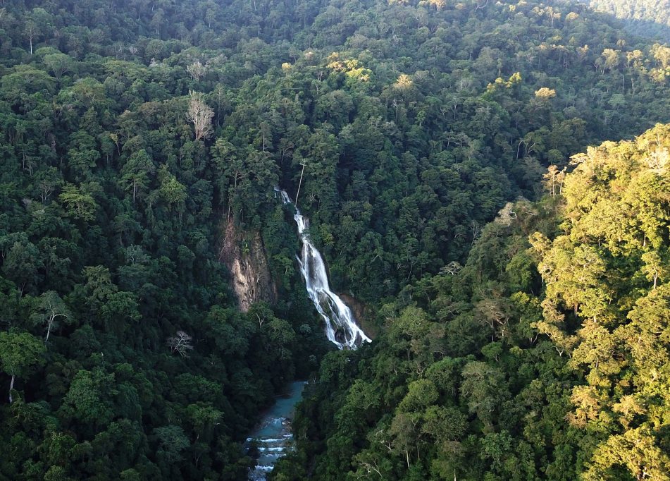 Lapopu Waterfall cascades down a steep, dark green forested cliff into a light blue river below, surrounded by dense tropical trees.