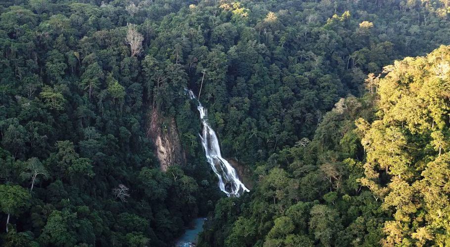Lapopu Waterfall cascades down a steep, dark green forested cliff into a light blue river below, surrounded by dense tropical trees.
