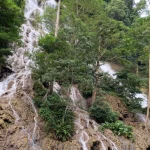 Lapopu Waterfall cascades down a multi-tiered, rocky cliff face covered in dense green foliage and trees under a bright sky.