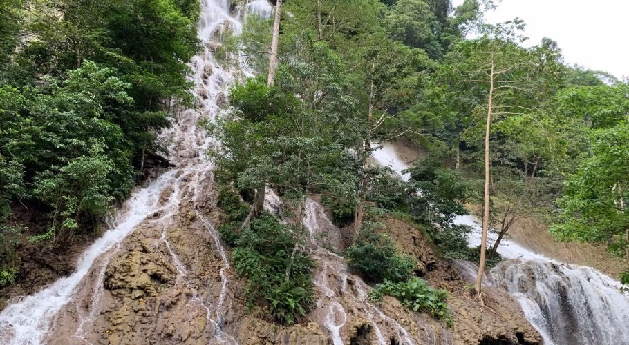 Lapopu Waterfall cascades down a multi-tiered, rocky cliff face covered in dense green foliage and trees under a bright sky.