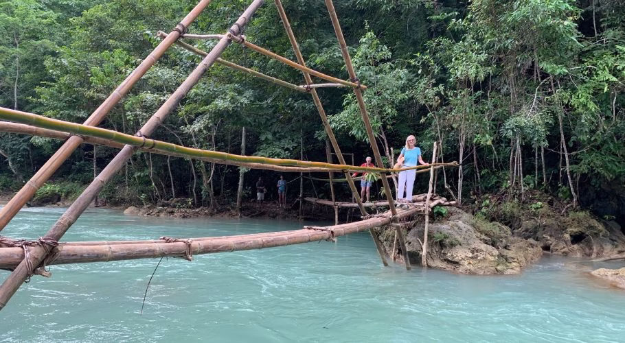 A woman in a blue top and white pants stands on a rustic bamboo bridge over a bright turquoise river, surrounded by lush green jungle.