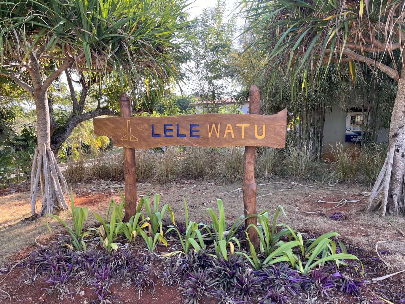 A carved wooden sign for "LELE WATU" stands amidst lush green and purple tropical plants on reddish-brown earth under a bright sky.
