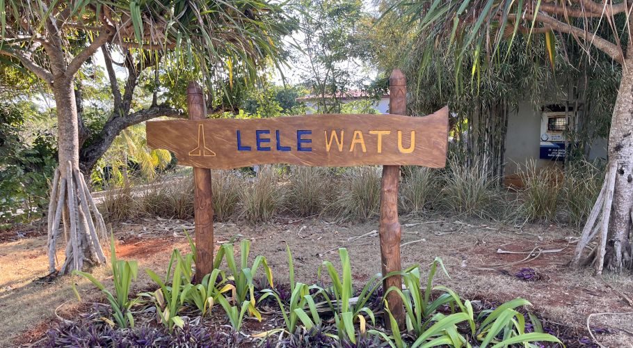 A carved wooden sign for "LELE WATU" stands amidst lush green and purple tropical plants on reddish-brown earth under a bright sky.