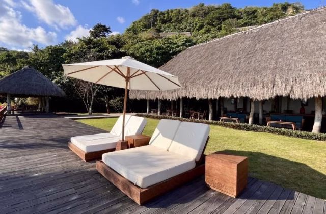 Two white sun loungers and a cream umbrella sit on a wooden deck beside a pool, facing a thatched-roof building and lush green hills under a blue sky.