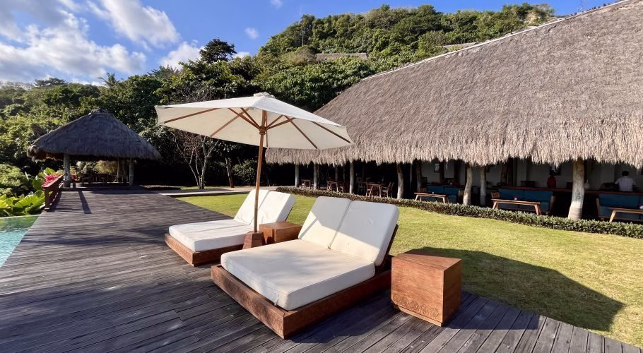 Two white sun loungers and a cream umbrella sit on a wooden deck beside a pool, facing a thatched-roof building and lush green hills under a blue sky.