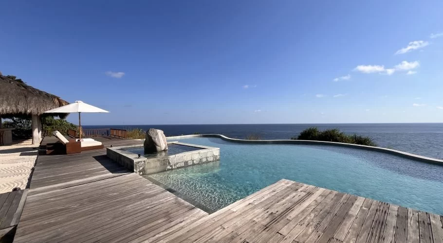 Light blue infinity pool and wooden deck overlook the dark blue ocean under a clear sky. A thatched cabana and lounge chair are left.