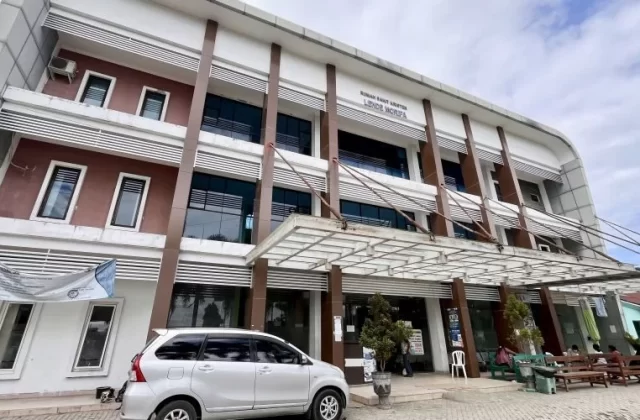 The modern Lende Moripa Hospital building in Waikabubak features white and brown facades, large dark windows, and an entrance canopy, with a silver car parked and people seated outside.