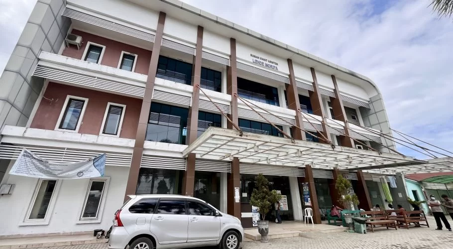 The modern Lende Moripa Hospital building in Waikabubak features white and brown facades, large dark windows, and an entrance canopy, with a silver car parked and people seated outside.