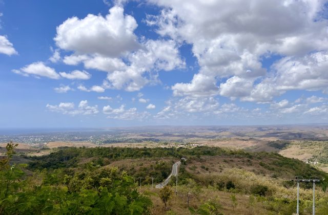 A panoramic view from a high viewpoint shows rolling hills with a winding road, a coastal city in the distance, and the ocean under a blue sky with white clouds.