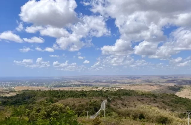 A panoramic view from a high viewpoint shows rolling hills with a winding road, a coastal city in the distance, and the ocean under a blue sky with white clouds.