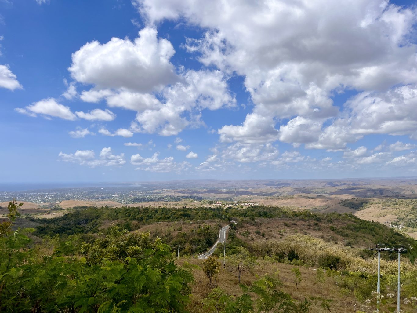 A panoramic view from a high viewpoint shows rolling hills with a winding road, a coastal city in the distance, and the ocean under a blue sky with white clouds.
