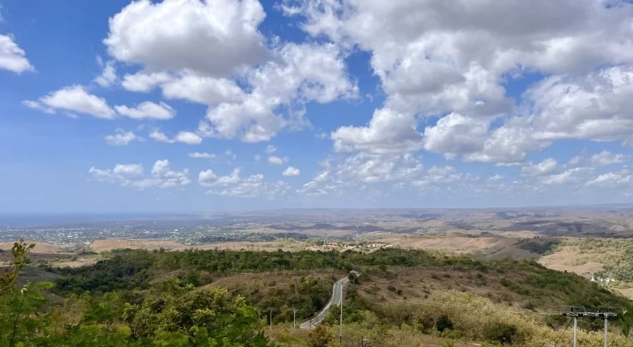 A panoramic view from a high viewpoint shows rolling hills with a winding road, a coastal city in the distance, and the ocean under a blue sky with white clouds.
