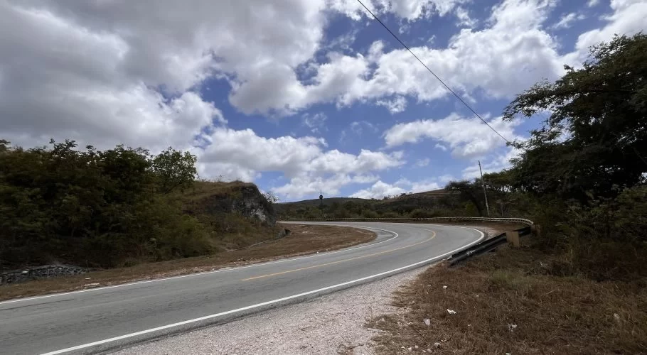 A winding paved road, known as the Letter S road, curves through dry, green hills under a partly cloudy blue sky in East Sumba.