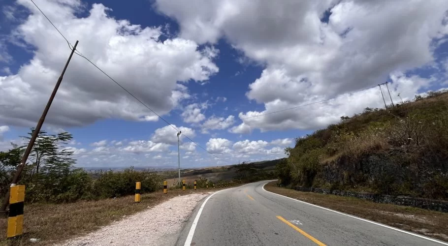 A winding asphalt road curves right, flanked by dry grassy hills and sparse trees under a blue sky with white clouds, offering a distant view of a city.