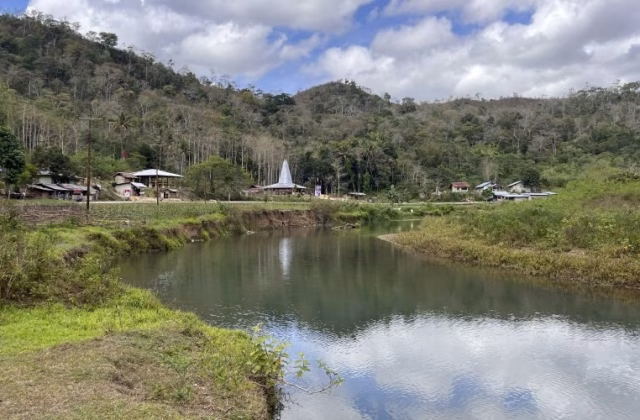 A serene river flows through a valley with green banks, a village featuring traditional Sumba houses, and a lush forested hill under a cloudy sky.