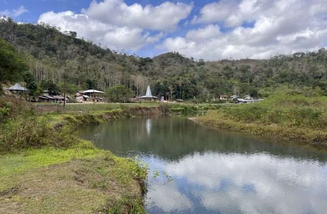 A serene river flows through a valley with green banks, a village featuring traditional Sumba houses, and a lush forested hill under a cloudy sky.