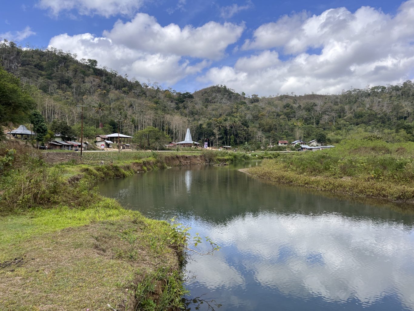 A serene river flows through a valley with green banks, a village featuring traditional Sumba houses, and a lush forested hill under a cloudy sky.