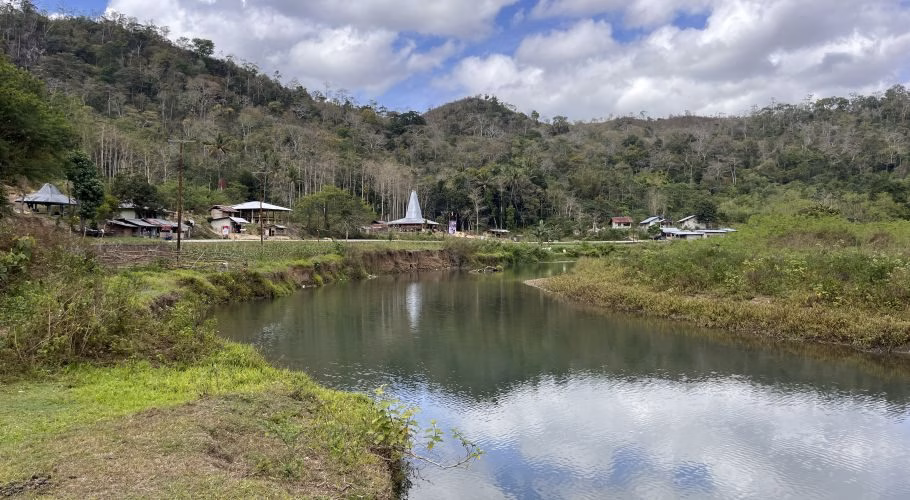 A serene river flows through a valley with green banks, a village featuring traditional Sumba houses, and a lush forested hill under a cloudy sky.