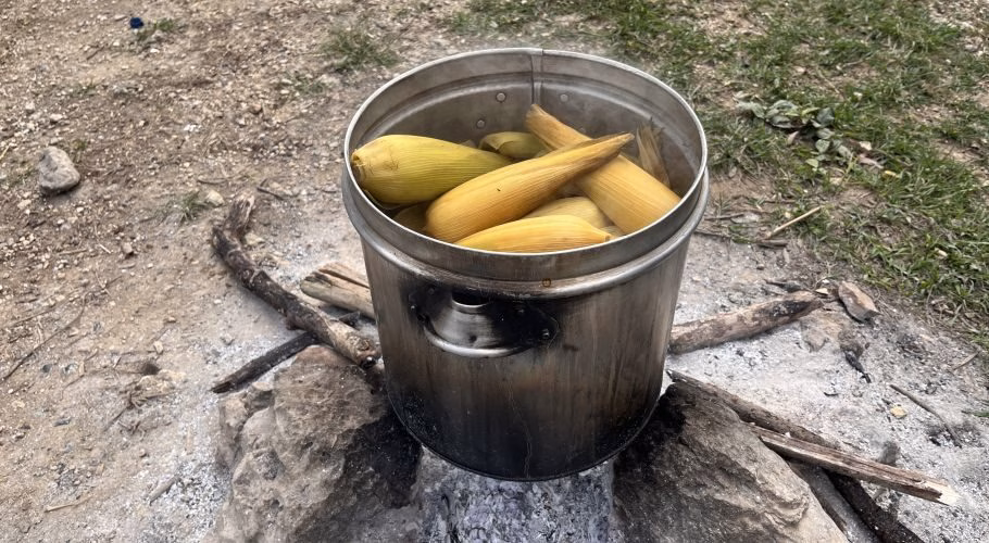 A large metal pot filled with yellow boiled corn cobs in husks steams over an outdoor fire pit on a dirt ground.