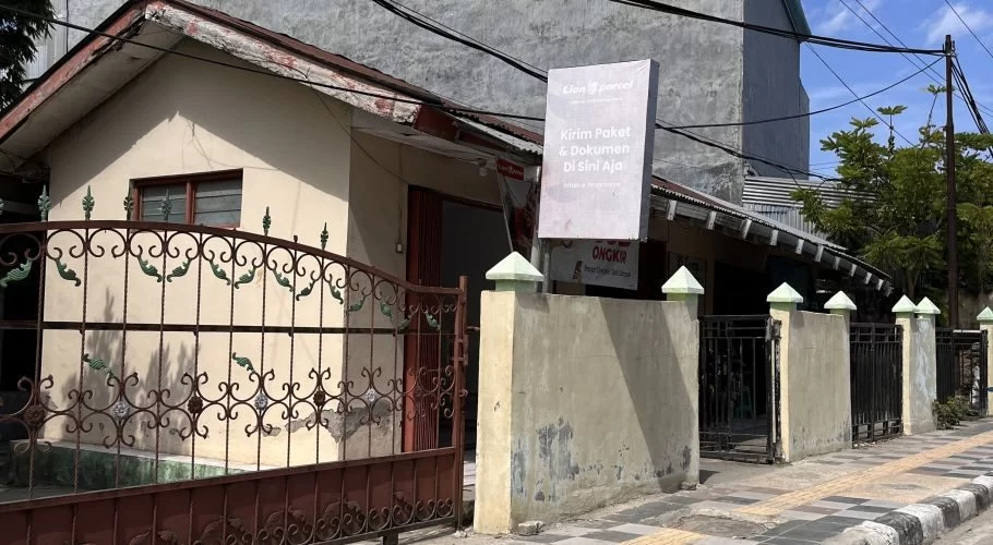 A light beige building with a brown ornate gate and a white 'Lion Parcel' sign stands by a sidewalk under a blue sky in Waikabubak, Sumba.