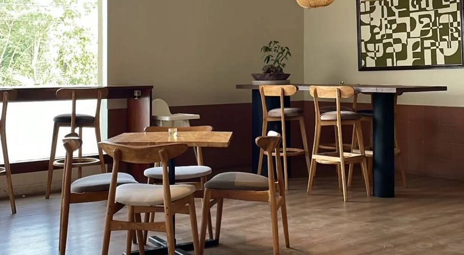 A modern cafe interior features light wooden tables and chairs, a two-toned wall, and an abstract green artwork, with a bright window showing outdoor greenery.