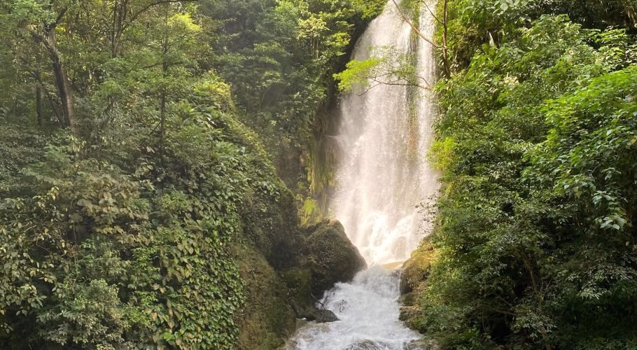 Lokomboro Waterfall plunges from a height, surrounded by dense green jungle foliage and mossy rocks, with water cascading into a pool.