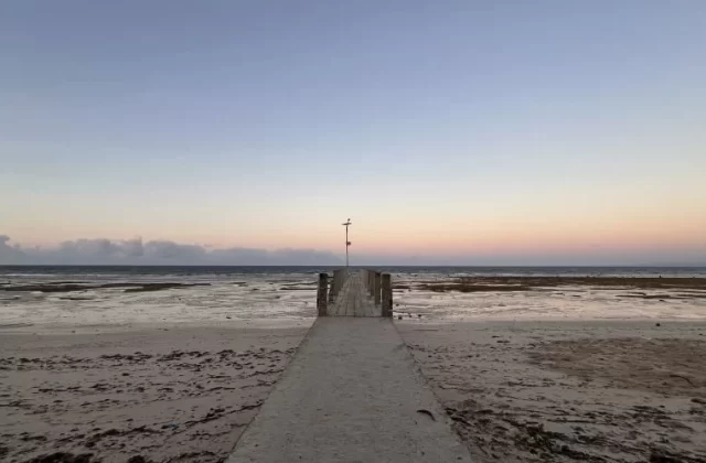A concrete path leads to a wooden pier extending into the shallow, calm ocean at Londa Lima Beach under a soft blue and pink twilight sky.