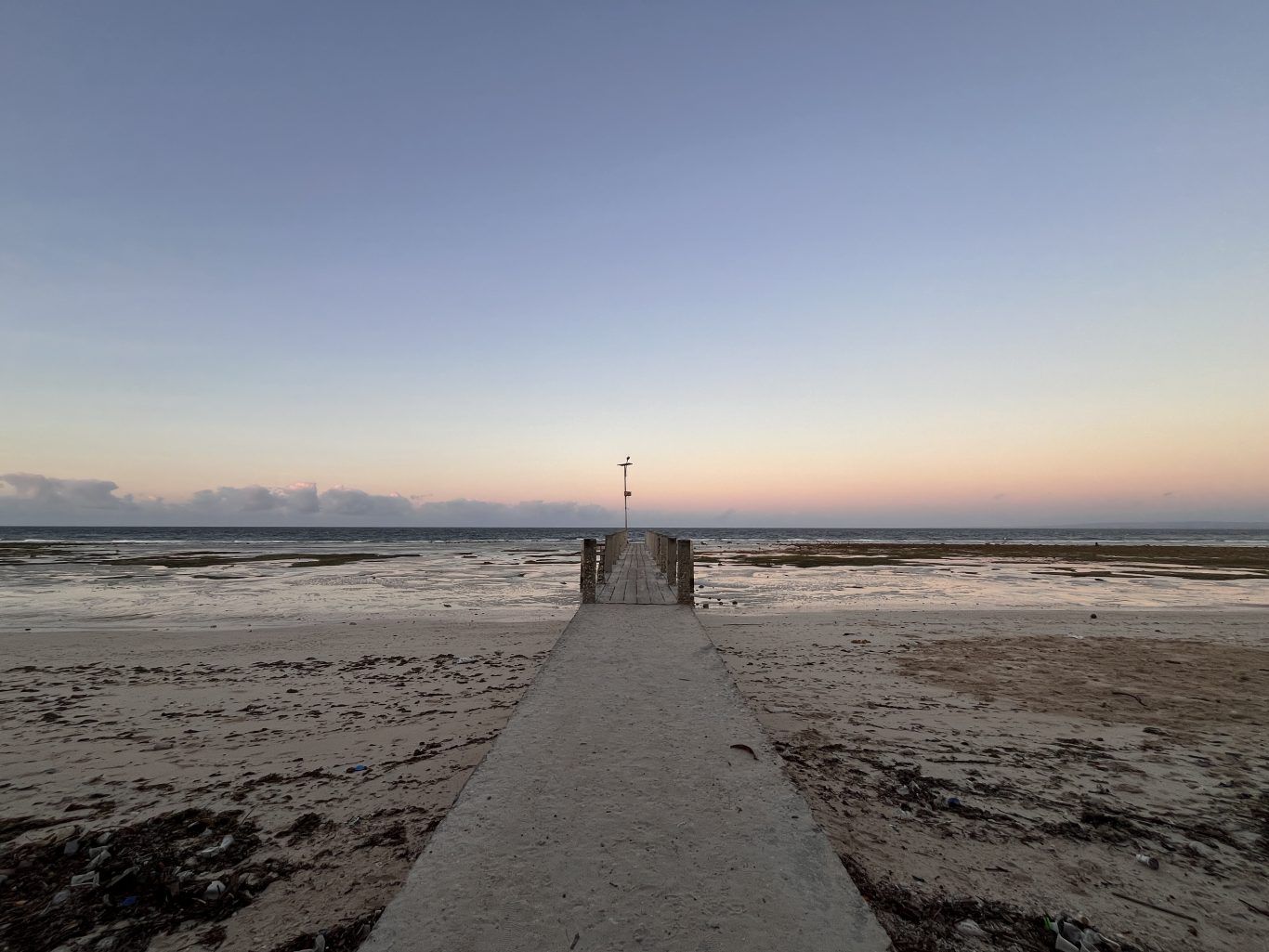 A concrete path leads to a wooden pier extending into the shallow, calm ocean at Londa Lima Beach under a soft blue and pink twilight sky.