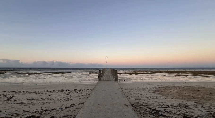 A concrete path leads to a wooden pier extending into the shallow, calm ocean at Londa Lima Beach under a soft blue and pink twilight sky.