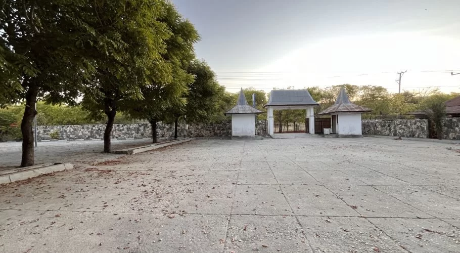 A wide, empty concrete plaza with green trees on the left and a traditional Indonesian entrance gate in the background, flanked by small white buildings with conical roofs, under a bright sky.