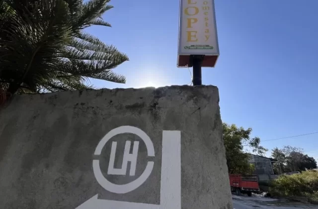 A concrete wall with a white "LH" logo and directional arrow points right towards the "LOPEY Homestay" sign under a bright blue sky.