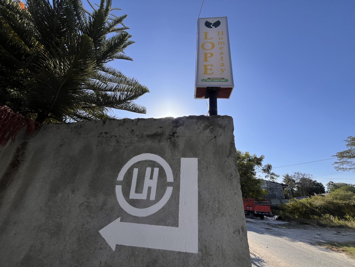 A concrete wall with a white "LH" logo and directional arrow points right towards the "LOPEY Homestay" sign under a bright blue sky.