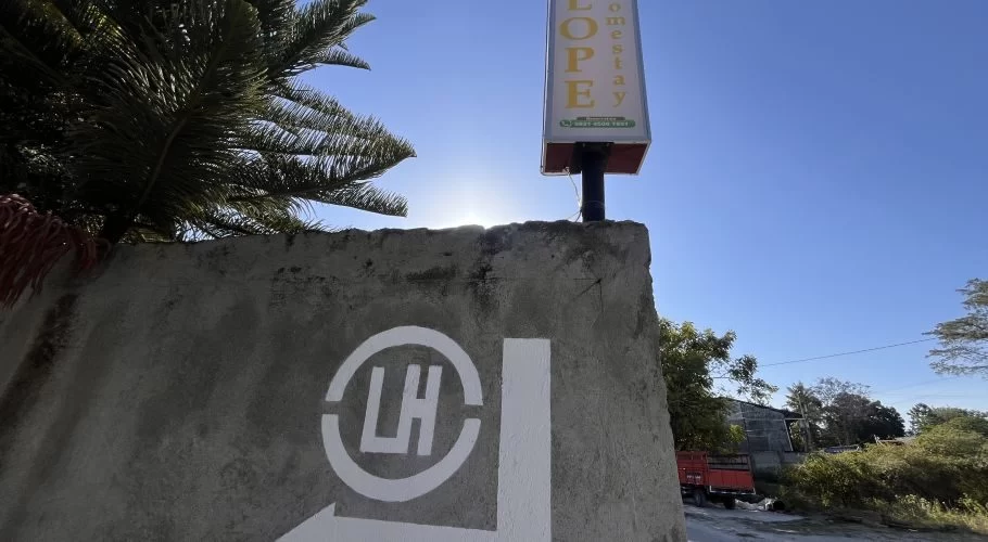 A concrete wall with a white "LH" logo and directional arrow points right towards the "LOPEY Homestay" sign under a bright blue sky.