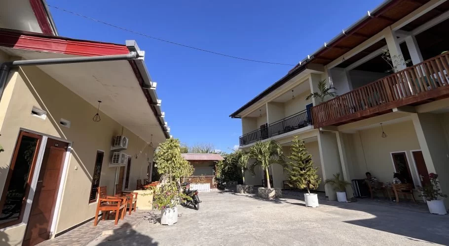 Two light-colored homestay buildings with red roofs face a paved courtyard, featuring outdoor seating, potted plants, and a clear blue sky.