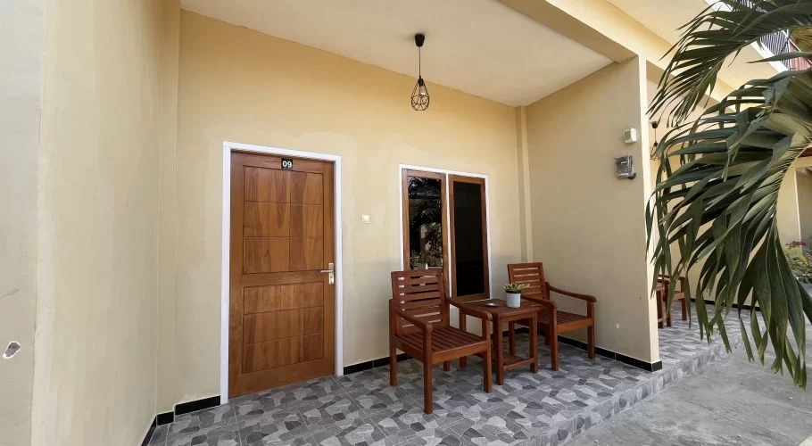 A light beige homestay room entrance with a brown wooden door, number 09, and a small patio featuring two wooden chairs and a table on grey patterned tiles.
