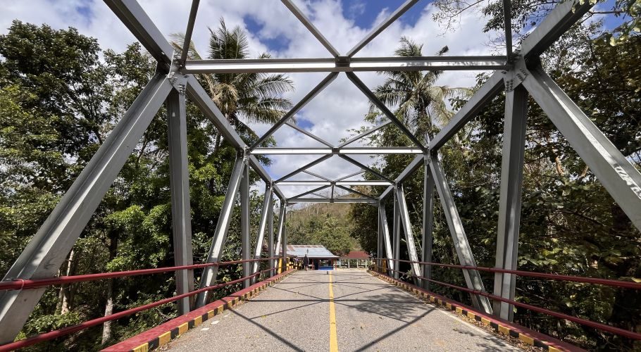 A grey metal truss bridge with red railings spans over a lush green forest under a partly cloudy blue sky, with a small building visible at its far end.