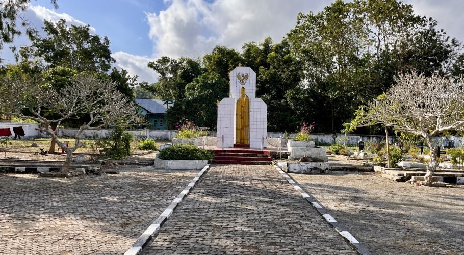 A paved path leads to the white Makam Pahlawan monument with a golden central column and an Indonesian eagle emblem, surrounded by trees.