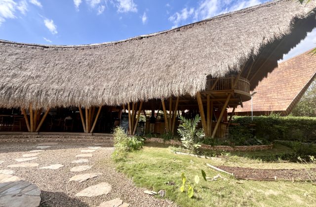 A large restaurant building with a wide, curved thatched roof supported by bamboo poles, featuring a second-floor balcony, a stone path, and green landscaping under a blue sky.
