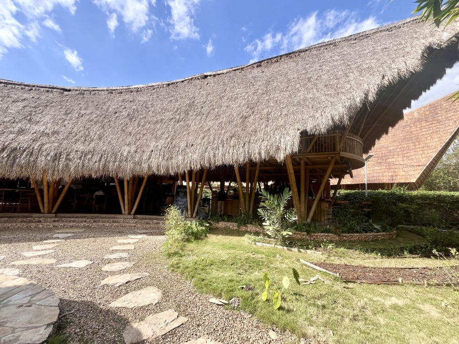 A large restaurant building with a wide, curved thatched roof supported by bamboo poles, featuring a second-floor balcony, a stone path, and green landscaping under a blue sky.