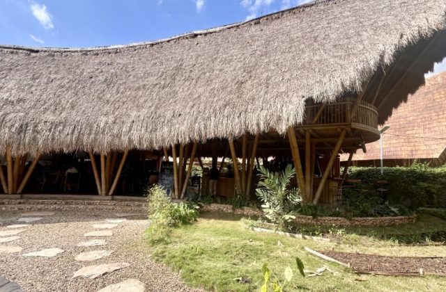 A large restaurant building with a wide, curved thatched roof supported by bamboo poles, featuring a second-floor balcony, a stone path, and green landscaping under a blue sky.