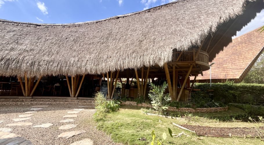 A large restaurant building with a wide, curved thatched roof supported by bamboo poles, featuring a second-floor balcony, a stone path, and green landscaping under a blue sky.