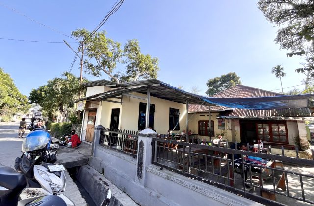 A cream-colored restaurant building with outdoor seating under a metal roof and blue tarp, a street with a scooter, and green trees under a clear blue sky in Waikabubak.