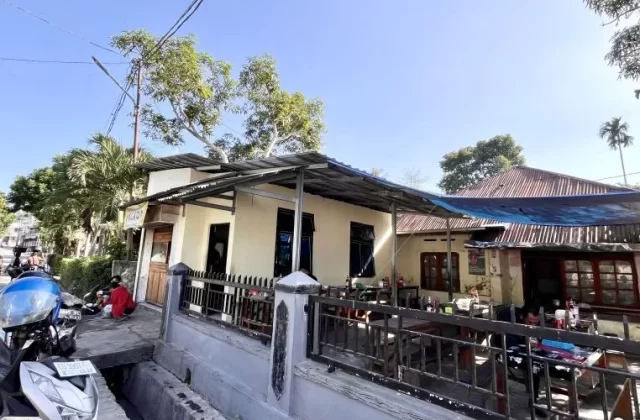 A cream-colored restaurant building with outdoor seating under a metal roof and blue tarp, a street with a scooter, and green trees under a clear blue sky in Waikabubak.