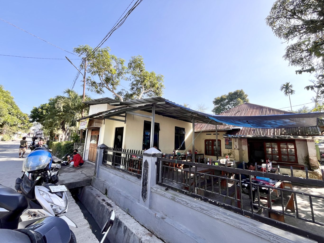 A cream-colored restaurant building with outdoor seating under a metal roof and blue tarp, a street with a scooter, and green trees under a clear blue sky in Waikabubak.
