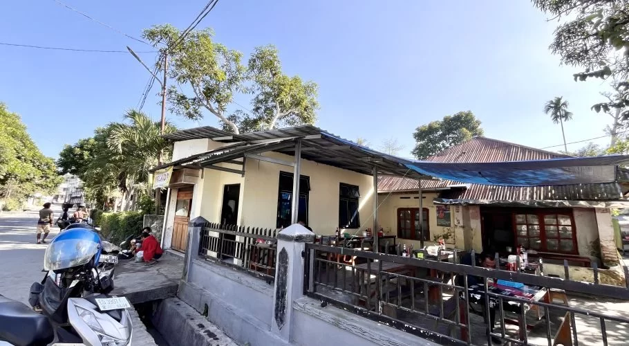 A cream-colored restaurant building with outdoor seating under a metal roof and blue tarp, a street with a scooter, and green trees under a clear blue sky in Waikabubak.