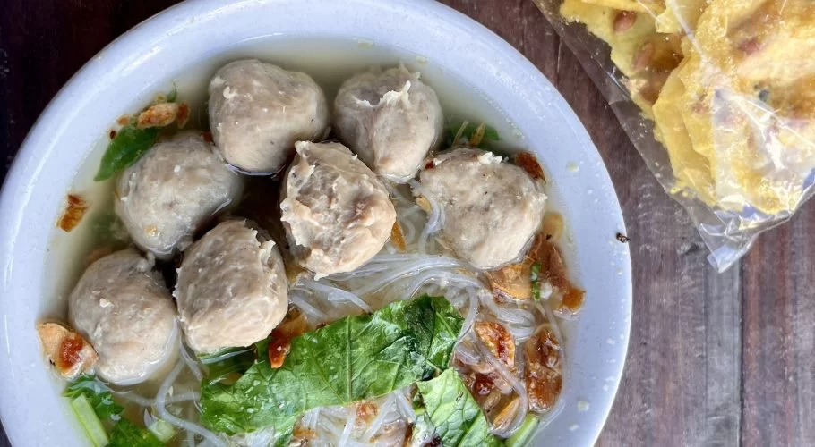 A white bowl of bakso soup with meatballs, noodles, green vegetables, and fried shallots, on a wooden table next to yellow crackers.