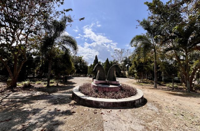 Two dark horse-head sculptures stand in a circular planter with reddish plants, surrounded by green trees and dirt paths under a blue, cloudy sky.