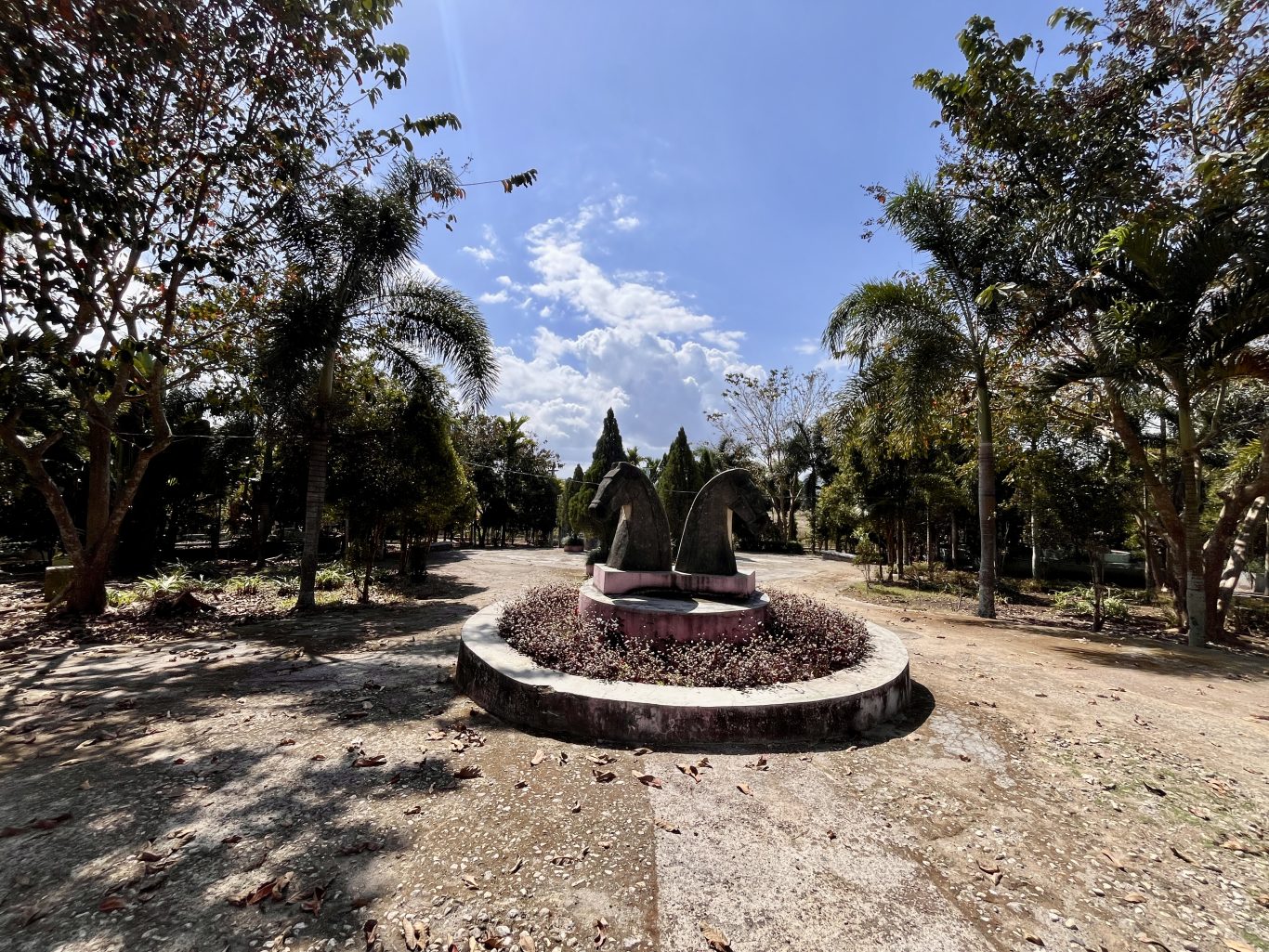 Two dark horse-head sculptures stand in a circular planter with reddish plants, surrounded by green trees and dirt paths under a blue, cloudy sky.